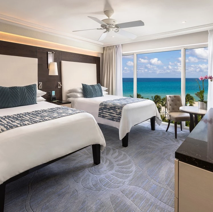 A wide-angle view of a hotel room with two beds, a large window overlooking the ocean, and modern decor.