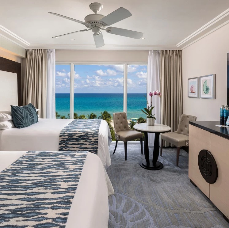 A hotel room with two beds, a table and chairs, and a large window overlooking the ocean.