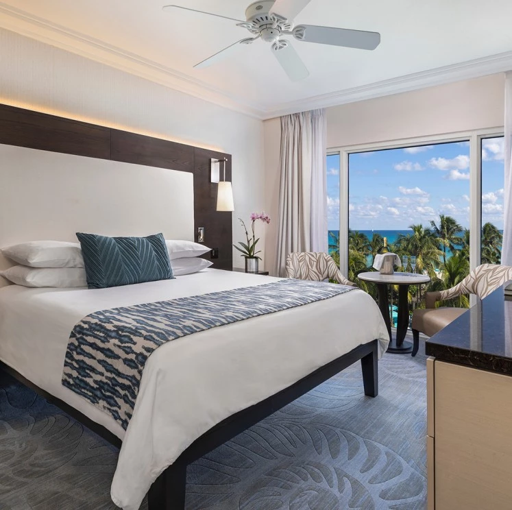 A wide-angle view of a hotel room with a large bed, sliding glass doors leading to a balcony with an ocean view, and a ceiling fan.