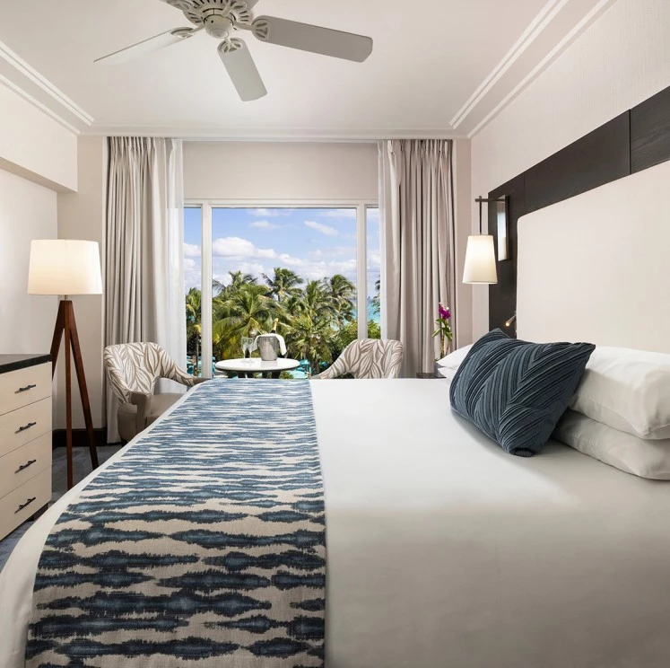 A wide-angle view of a hotel room with a large bed, a window with a view of palm trees, and a seating area.