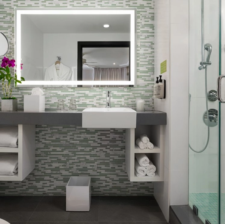 A modern bathroom with a large led light mirror, glass shower door, tiled backsplash, floating sink, and built-in shelves.