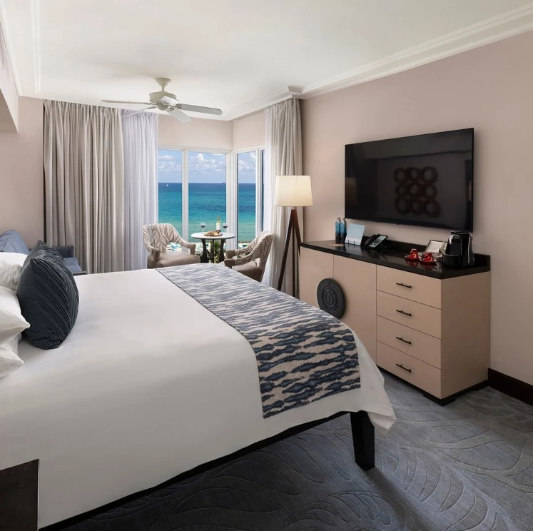 A hotel room with a bed, dresser, TV, and chair in front of a window with an ocean view.