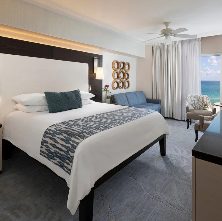 A wide-angle view of a hotel room with a large bed, sofa, chair, and desk, featuring an ocean view.