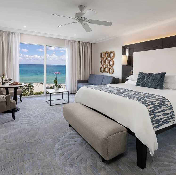 A wide-angle view of a hotel room with a large bed, sofa, coffee table, dining table, and ocean view.