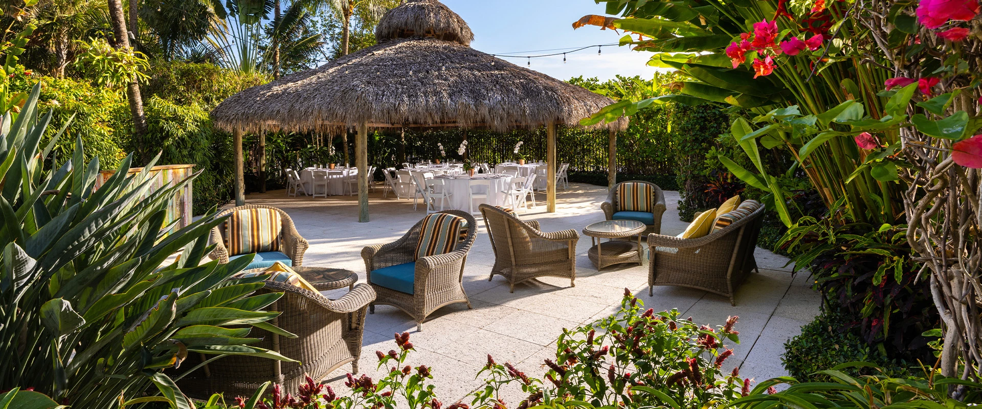 A tropical garden seating area with wicker furniture and a thatched roof structure.