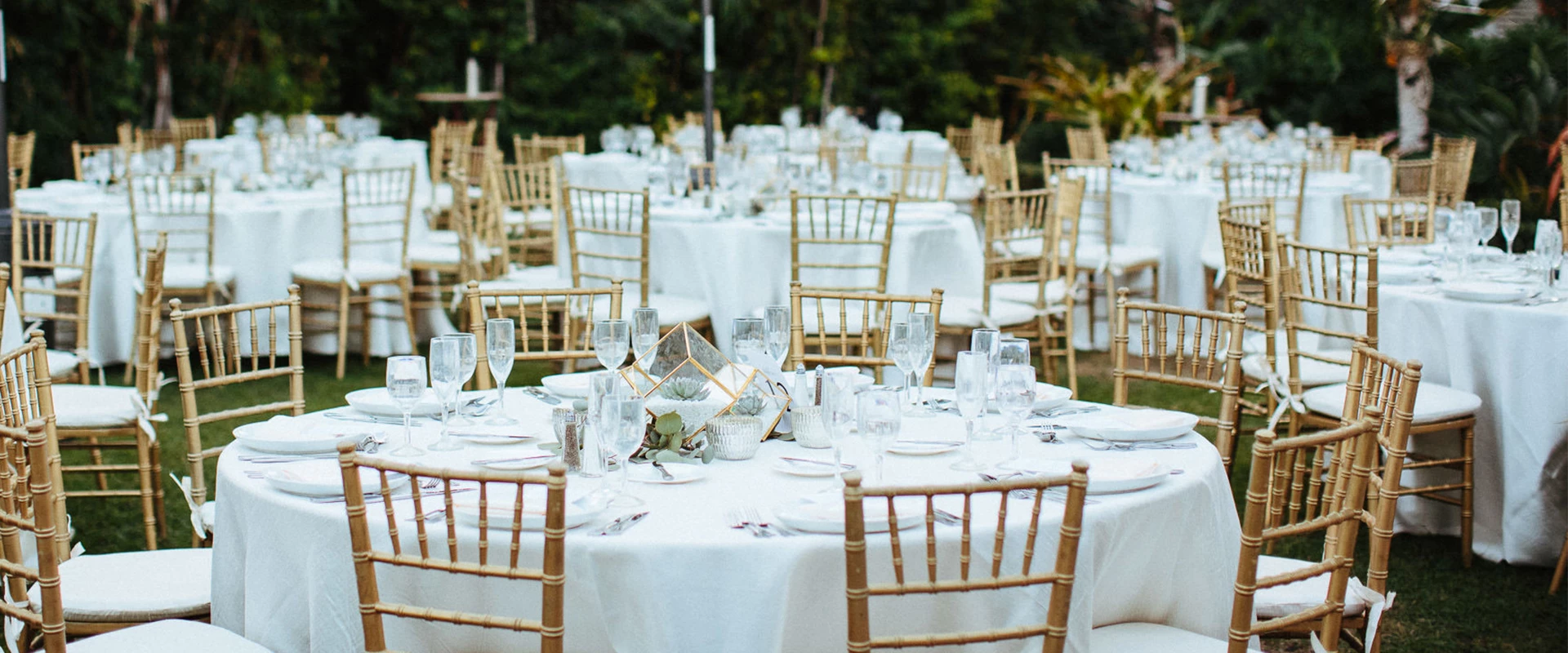 A series of round tables with white tablecloths, gold chairs, and elegant centerpieces set up for an outdoor event.