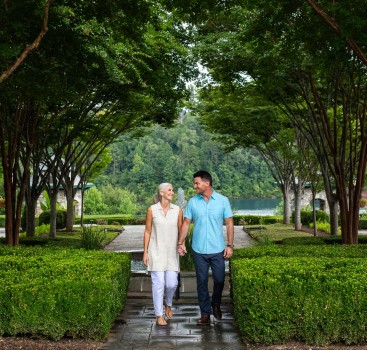a man and woman holding hands walking on a path with trees and water
