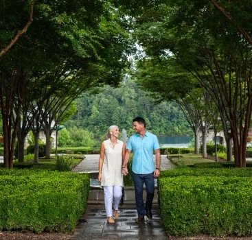 a man and woman holding hands walking on a path with trees and water