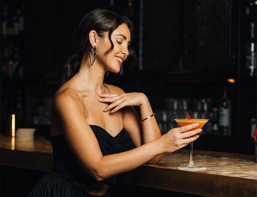 a girl wearing a black dress holding an orange colored martini while standing at a low lit bar