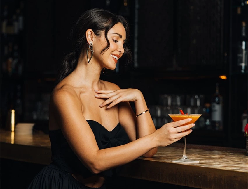 a girl wearing a black dress holding an orange colored martini while standing at a low lit bar
