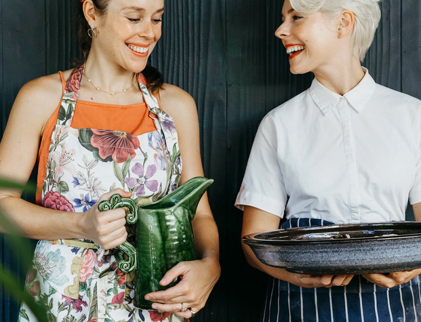 2 woman smiling at each other holding pitcher and plate