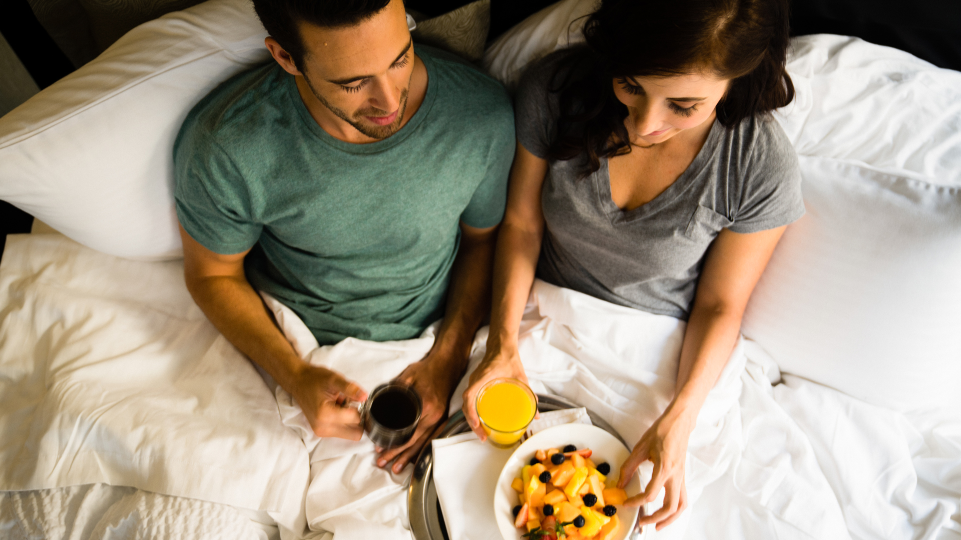 a man and woman sitting in bed with a plate of fruit and juice