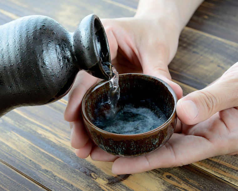 drink being poured from a ceramic bottle into a cup that a person is holding 