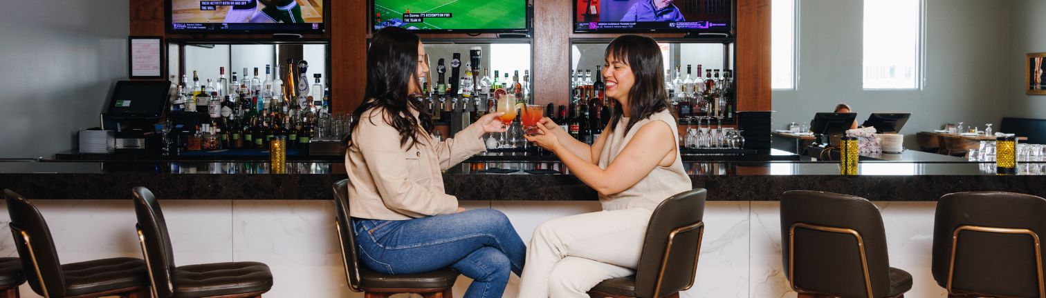 two women sitting at a bar toasting with drinks