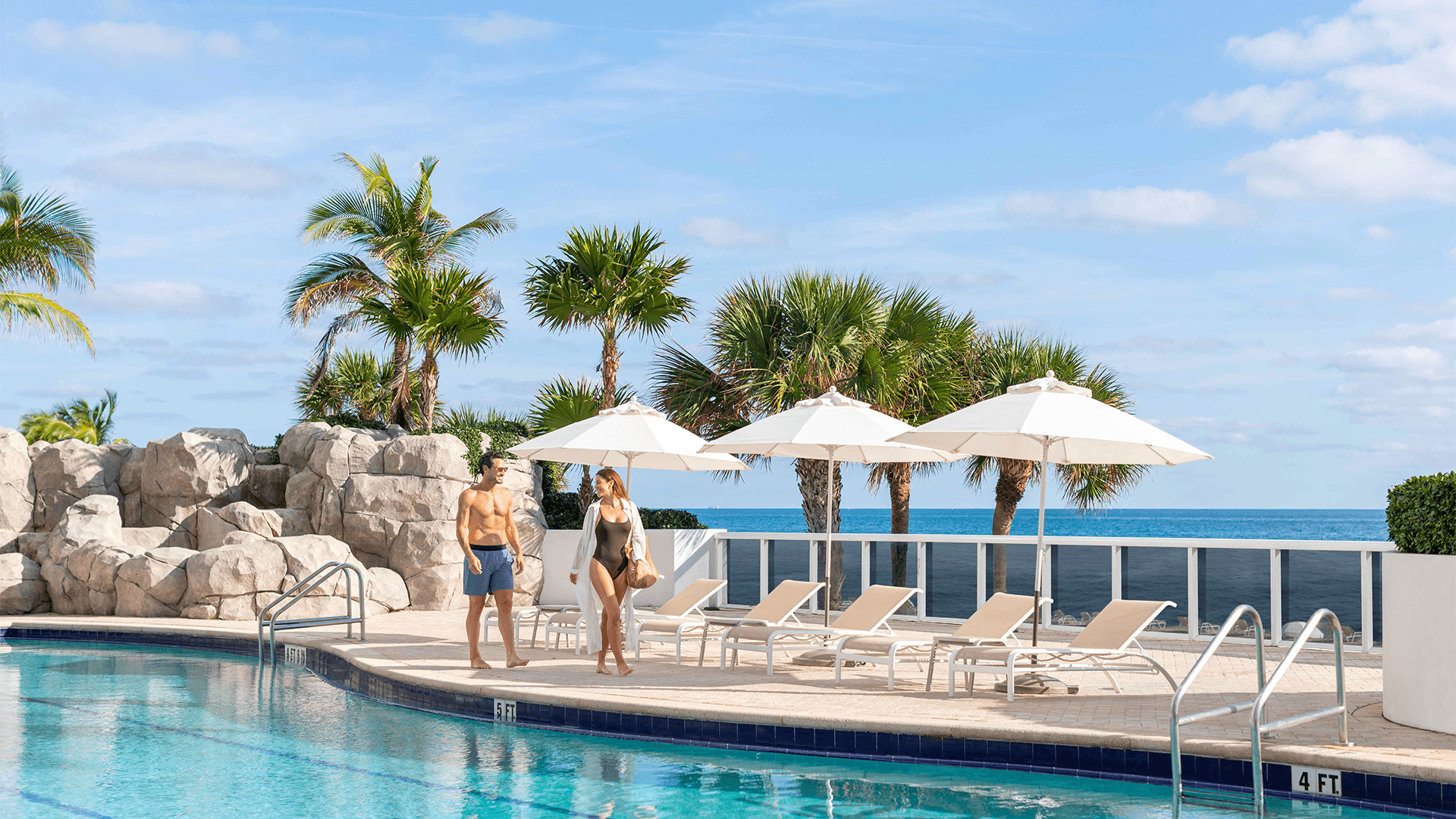 a man and woman standing next to a pool with umbrellas and palm trees