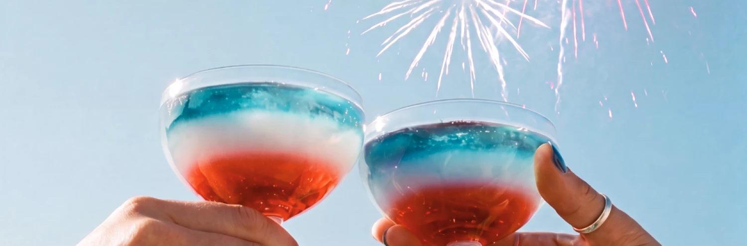 two hands holding glasses with red and blue liquid and fireworks in the background