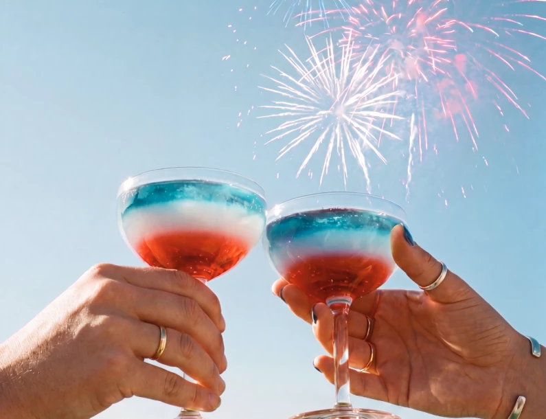 two hands holding glasses with red and blue liquid and fireworks in the background