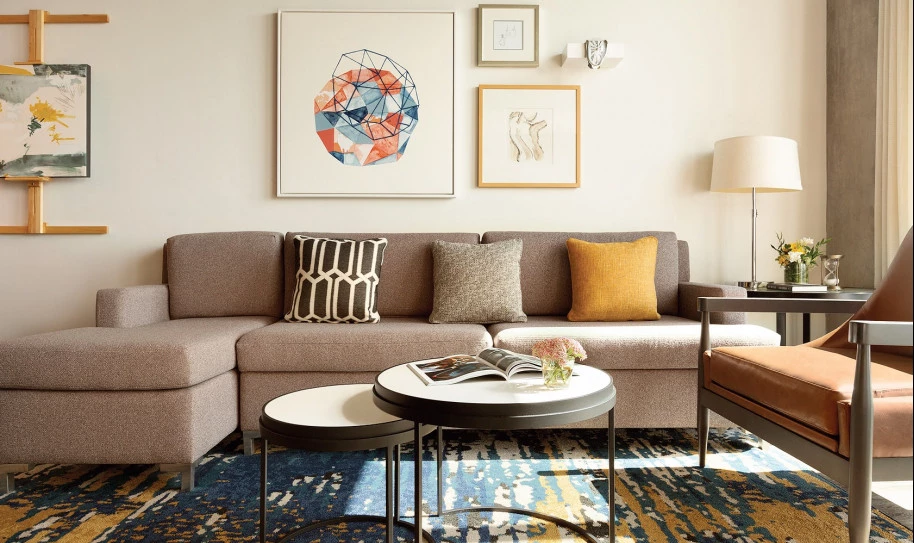 A gray couch with throw pillows, one yellow and one black with white patterns, is in a living room with a circular coffee table, various paintings on the walls, wood floors, a designer carpet, and two hightop chairs angled against a kitchen bar