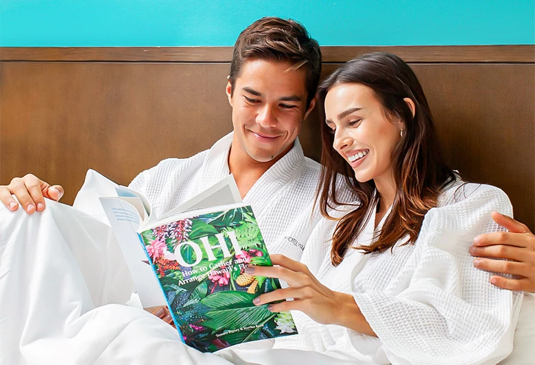 A man and woman in white robes sit on a bed, reading a magazine.