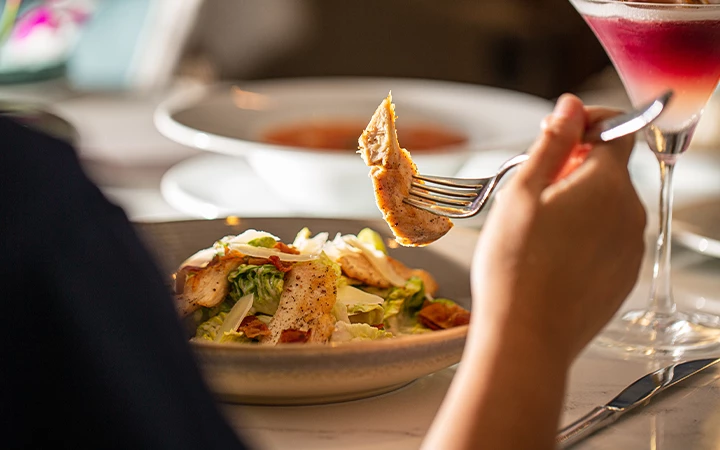 A hand holding a fork with a piece of food over a plate of salad and a glass of red drink.