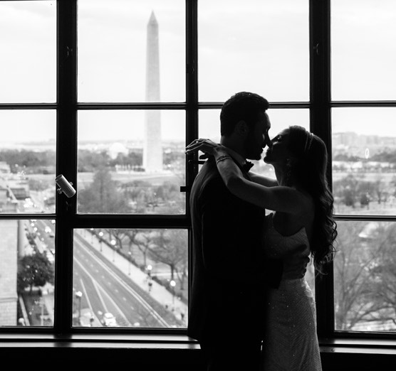 black and white photo of bride and groom almost kissing in front of window with washington monument in the background