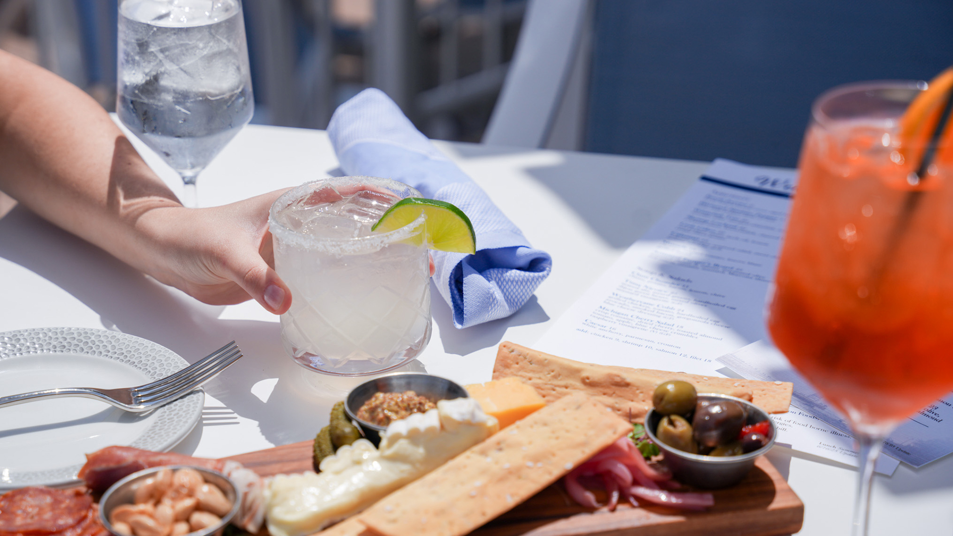 a hand holding a drink with a lime wedge and a plate of food