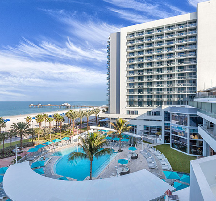 view of hotel pool area with hotel and beach in background on a sunny day