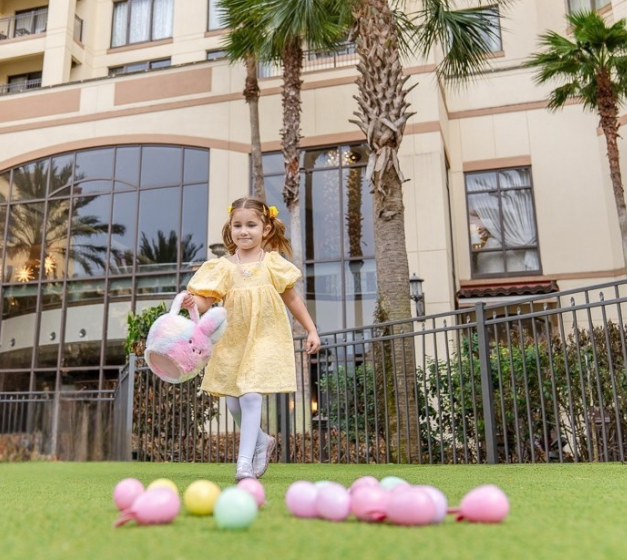 a girl in a yellow dress walking on grass with a pink bunny basket and eggs