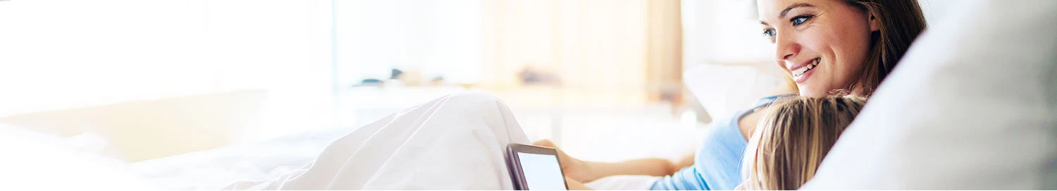 Woman and child laying in bet with soft pillowy linens looking at a tablet computer