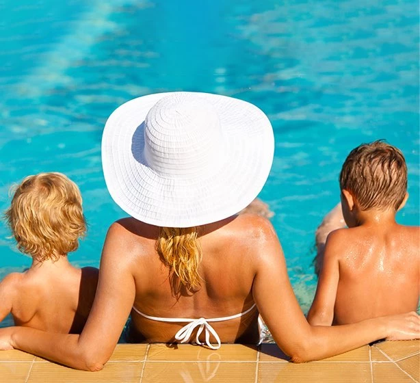 children and woman lounging poolside wearing a white swimsuit and white sun hat