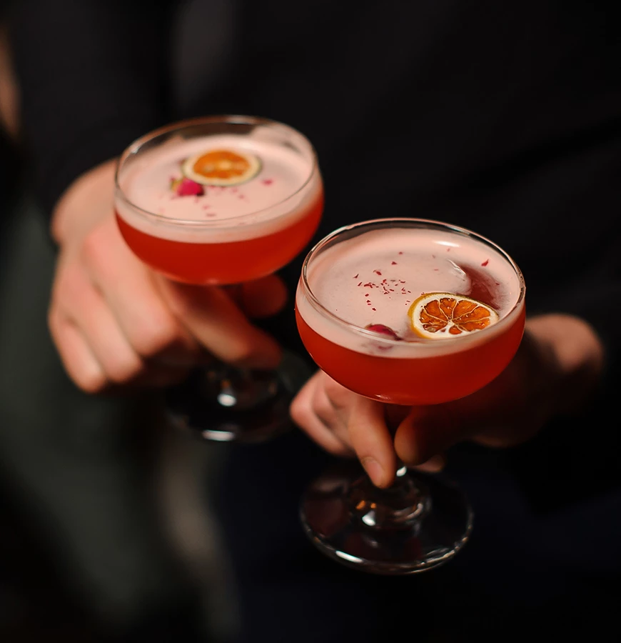 a pair of hands holding glasses with pink liquid and orange slices