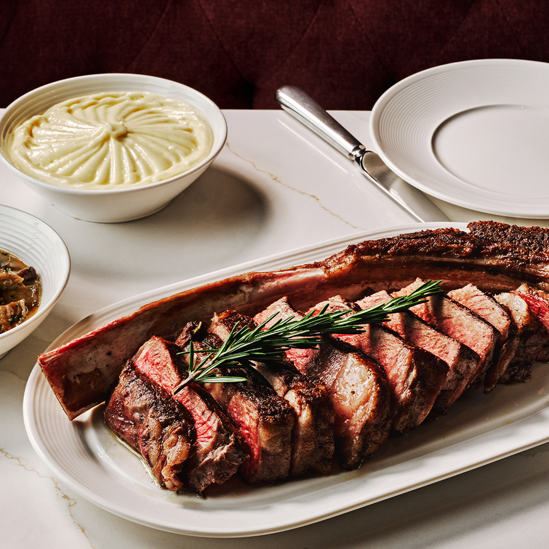 a plate of meat with a rosemary sprig on it next to bowls of mashed potatoes